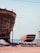 Two large boats are situated on a sandy shore near an ocean. The boat in the foreground is unfinished, revealing its wooden structure and scaffolding. The second boat in the background is painted red and white, resting on supports. Several blue barrels and construction materials are scattered around, indicating a boat-building or repair site.