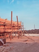 A large wooden structure under construction is supported by numerous beams and planks. The setting appears to be a construction site with visible mountains in the background and a tall electrical tower on the right. The foreground has some scattered construction materials on the ground.