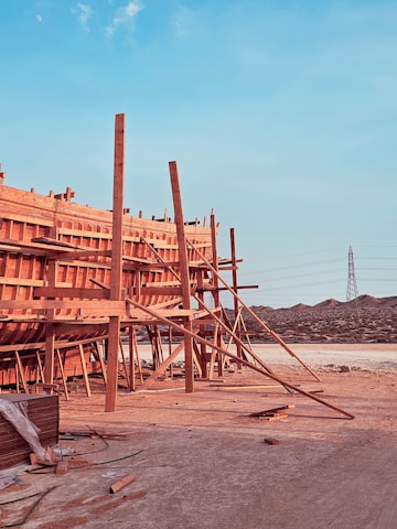 A large wooden structure under construction is supported by numerous beams and planks. The setting appears to be a construction site with visible mountains in the background and a tall electrical tower on the right. The foreground has some scattered construction materials on the ground.
