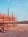 A large wooden structure under construction is supported by numerous beams and planks. The setting appears to be a construction site with visible mountains in the background and a tall electrical tower on the right. The foreground has some scattered construction materials on the ground.