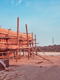 A large wooden structure under construction is supported by numerous beams and planks. The setting appears to be a construction site with visible mountains in the background and a tall electrical tower on the right. The foreground has some scattered construction materials on the ground.