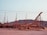 Wooden structures with scaffolding are set up against a backdrop of rocky hills under a clear sky. The framework appears to be part of a large construction project, possibly a ship or building, with beams and supports in place.