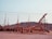 Wooden structures with scaffolding are set up against a backdrop of rocky hills under a clear sky. The framework appears to be part of a large construction project, possibly a ship or building, with beams and supports in place.