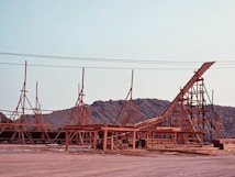 Wooden structures with scaffolding are set up against a backdrop of rocky hills under a clear sky. The framework appears to be part of a large construction project, possibly a ship or building, with beams and supports in place.