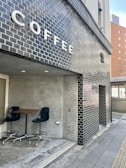 A modern coffee shop exterior is seen with black brick walls and a large sign spelling out 'COFFEE' in bold white letters. A small seating area is visible, featuring two dark blue chairs and a wooden table against a gray concrete wall. The setting gives a clean and minimalist urban vibe.