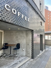 A modern coffee shop exterior is seen with black brick walls and a large sign spelling out 'COFFEE' in bold white letters. A small seating area is visible, featuring two dark blue chairs and a wooden table against a gray concrete wall. The setting gives a clean and minimalist urban vibe.