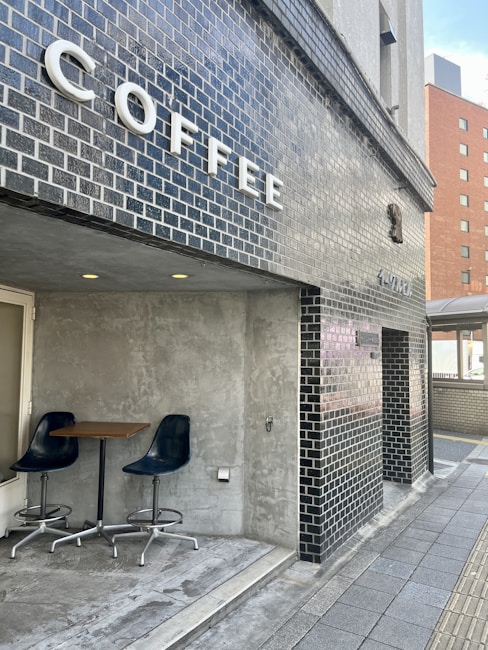 A modern coffee shop exterior is seen with black brick walls and a large sign spelling out 'COFFEE' in bold white letters. A small seating area is visible, featuring two dark blue chairs and a wooden table against a gray concrete wall. The setting gives a clean and minimalist urban vibe.