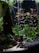 An aquarium scene featuring lush green aquatic plants and small orange fish swimming among the foliage. The base of the aquarium is lined with dark rocks and sandy substrate. The plants provide a dense, natural look with varying shades of green and hints of red.