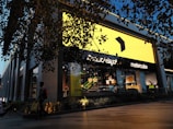 A modern retail store with a large bright yellow sign dominates the scene. The storefront is stylishly decorated with contemporary furniture visible through the glass. A person carrying a yellow shopping bag walks past, and there is a promotional sign offering up to 50% off. The store is surrounded by greenery, and overhead branches add to the ambiance. Evening light casts a warm glow, enhancing the visual appeal.