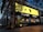 A modern retail store with a large bright yellow sign dominates the scene. The storefront is stylishly decorated with contemporary furniture visible through the glass. A person carrying a yellow shopping bag walks past, and there is a promotional sign offering up to 50% off. The store is surrounded by greenery, and overhead branches add to the ambiance. Evening light casts a warm glow, enhancing the visual appeal.