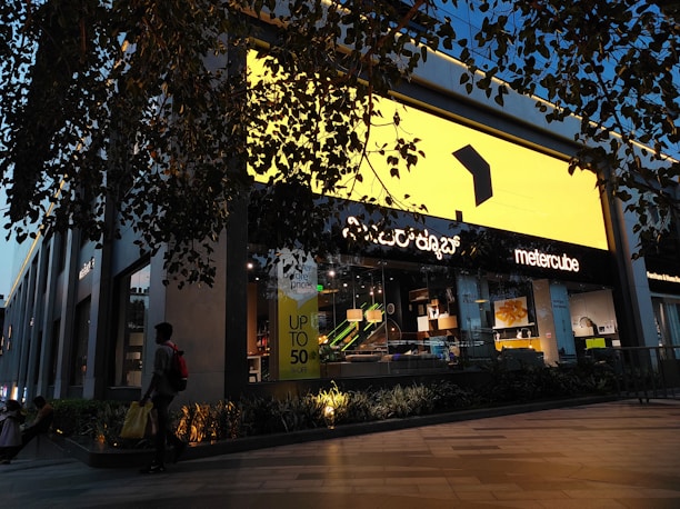 A modern retail store with a large bright yellow sign dominates the scene. The storefront is stylishly decorated with contemporary furniture visible through the glass. A person carrying a yellow shopping bag walks past, and there is a promotional sign offering up to 50% off. The store is surrounded by greenery, and overhead branches add to the ambiance. Evening light casts a warm glow, enhancing the visual appeal.