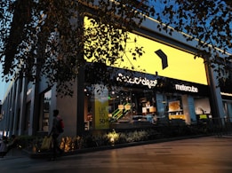 A modern retail store with a large bright yellow sign dominates the scene. The storefront is stylishly decorated with contemporary furniture visible through the glass. A person carrying a yellow shopping bag walks past, and there is a promotional sign offering up to 50% off. The store is surrounded by greenery, and overhead branches add to the ambiance. Evening light casts a warm glow, enhancing the visual appeal.