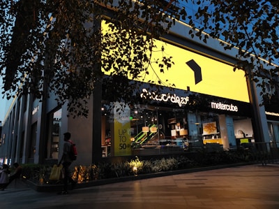A modern retail store with a large bright yellow sign dominates the scene. The storefront is stylishly decorated with contemporary furniture visible through the glass. A person carrying a yellow shopping bag walks past, and there is a promotional sign offering up to 50% off. The store is surrounded by greenery, and overhead branches add to the ambiance. Evening light casts a warm glow, enhancing the visual appeal.