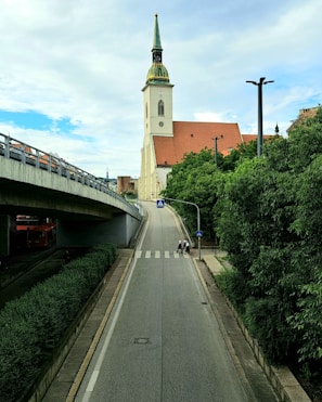 A tall church with a distinctive green and gold spire stands prominently against a partly cloudy sky. The church is situated near a wide, sloping road with a pedestrian crossing marked by two signs. On the left, there is an overpass with vehicles, and lush green trees line the pathway on the right side.