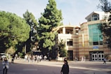 A university campus scene with a modern building featuring large glass windows and a sign that says 'UCLA Store'. In the foreground, people walk along a plaza lined with tall green trees. The overall atmosphere is bustling with students and visitors.