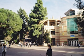 A university campus scene with a modern building featuring large glass windows and a sign that says 'UCLA Store'. In the foreground, people walk along a plaza lined with tall green trees. The overall atmosphere is bustling with students and visitors.