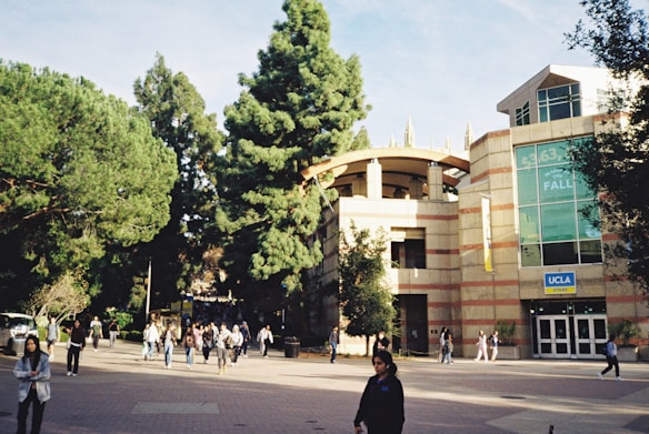 A university campus scene with a modern building featuring large glass windows and a sign that says 'UCLA Store'. In the foreground, people walk along a plaza lined with tall green trees. The overall atmosphere is bustling with students and visitors.