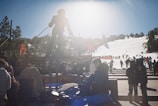 A bright winter day at a ski resort with a statue of a skier silhouetted against the sun. Visitors and skiers are gathered around, with many preparing near the base and slopes in the background. The area is bustling with activity.