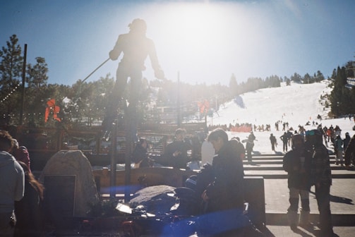 A bright winter day at a ski resort with a statue of a skier silhouetted against the sun. Visitors and skiers are gathered around, with many preparing near the base and slopes in the background. The area is bustling with activity.