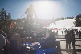 A bright winter day at a ski resort with a statue of a skier silhouetted against the sun. Visitors and skiers are gathered around, with many preparing near the base and slopes in the background. The area is bustling with activity.