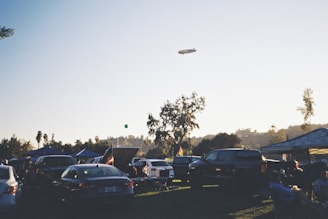 A parking area filled with numerous cars under a clear sky during early evening. Several canopies are set up, likely for a gathering or event. A blimp is seen floating in the sky. People are sitting and standing around the parked vehicles, suggesting a relaxed, social atmosphere. In the background, trees are silhouetted against the setting sun.