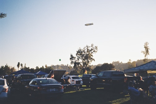 A parking area filled with numerous cars under a clear sky during early evening. Several canopies are set up, likely for a gathering or event. A blimp is seen floating in the sky. People are sitting and standing around the parked vehicles, suggesting a relaxed, social atmosphere. In the background, trees are silhouetted against the setting sun.