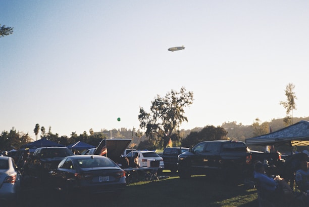 A parking area filled with numerous cars under a clear sky during early evening. Several canopies are set up, likely for a gathering or event. A blimp is seen floating in the sky. People are sitting and standing around the parked vehicles, suggesting a relaxed, social atmosphere. In the background, trees are silhouetted against the setting sun.
