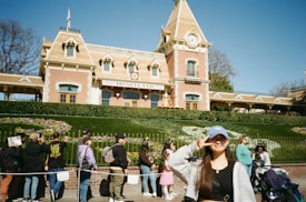 A young woman is posing for a photo in front of a Disneyland entrance, with a crowd of people in the background. The building is an ornate, brick structure with a clock tower and is surrounded by well-manicured gardens featuring flowers. The sky is clear and blue, suggesting a sunny day.