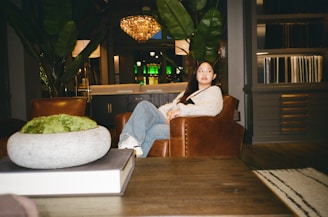 A person sits in a cozy, well-decorated living room with a modern yet rustic style. The room features a large chandelier, large leafy plants, a dark wooden coffee table, and leather armchairs. The setting is warm and inviting, with ambient lighting and decorative elements like books and a moss centerpiece on the table.