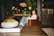 A person sits in a cozy, well-decorated living room with a modern yet rustic style. The room features a large chandelier, large leafy plants, a dark wooden coffee table, and leather armchairs. The setting is warm and inviting, with ambient lighting and decorative elements like books and a moss centerpiece on the table.