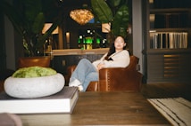 A person sits in a cozy, well-decorated living room with a modern yet rustic style. The room features a large chandelier, large leafy plants, a dark wooden coffee table, and leather armchairs. The setting is warm and inviting, with ambient lighting and decorative elements like books and a moss centerpiece on the table.