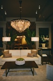 A luxurious living room features a large, elegant chandelier casting a warm glow over the space. A plush cream sofa with several earthy throw pillows faces a dark wooden coffee table adorned with a decorative potted plant. Two tall lamps flank the sofa, and large green plants add a touch of nature. The room is bordered by mirrors and understated decor, creating a sophisticated and inviting ambiance.