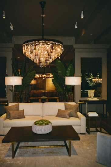 A luxurious living room features a large, elegant chandelier casting a warm glow over the space. A plush cream sofa with several earthy throw pillows faces a dark wooden coffee table adorned with a decorative potted plant. Two tall lamps flank the sofa, and large green plants add a touch of nature. The room is bordered by mirrors and understated decor, creating a sophisticated and inviting ambiance.