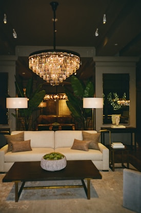 A luxurious living room features a large, elegant chandelier casting a warm glow over the space. A plush cream sofa with several earthy throw pillows faces a dark wooden coffee table adorned with a decorative potted plant. Two tall lamps flank the sofa, and large green plants add a touch of nature. The room is bordered by mirrors and understated decor, creating a sophisticated and inviting ambiance.