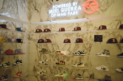 A display wall features a collection of baseball caps and sneakers arranged on transparent shelves against a marble backdrop. The caps exhibit various team logos, while the sneakers include different styles and colorations. A neon sign above the display reads 'Powered by Miki Guerra of Mag Park.'