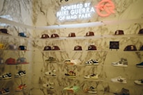 A display wall features a collection of baseball caps and sneakers arranged on transparent shelves against a marble backdrop. The caps exhibit various team logos, while the sneakers include different styles and colorations. A neon sign above the display reads 'Powered by Miki Guerra of Mag Park.'