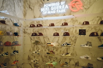 A display wall features a collection of baseball caps and sneakers arranged on transparent shelves against a marble backdrop. The caps exhibit various team logos, while the sneakers include different styles and colorations. A neon sign above the display reads 'Powered by Miki Guerra of Mag Park.'