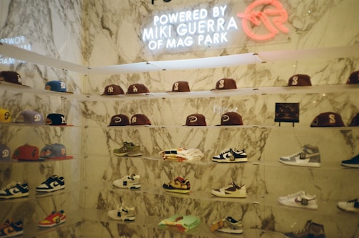 A display wall features a collection of baseball caps and sneakers arranged on transparent shelves against a marble backdrop. The caps exhibit various team logos, while the sneakers include different styles and colorations. A neon sign above the display reads 'Powered by Miki Guerra of Mag Park.'