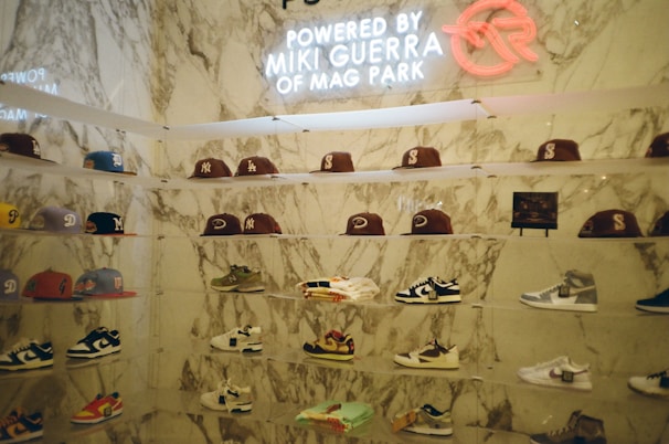 A display wall features a collection of baseball caps and sneakers arranged on transparent shelves against a marble backdrop. The caps exhibit various team logos, while the sneakers include different styles and colorations. A neon sign above the display reads 'Powered by Miki Guerra of Mag Park.'
