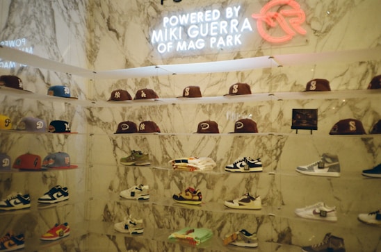A display wall features a collection of baseball caps and sneakers arranged on transparent shelves against a marble backdrop. The caps exhibit various team logos, while the sneakers include different styles and colorations. A neon sign above the display reads 'Powered by Miki Guerra of Mag Park.'