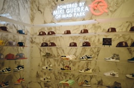 A display wall features a collection of baseball caps and sneakers arranged on transparent shelves against a marble backdrop. The caps exhibit various team logos, while the sneakers include different styles and colorations. A neon sign above the display reads 'Powered by Miki Guerra of Mag Park.'
