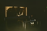 A person sits on a bench in a dimly lit room, observing a large, abstract, monochromatic artwork displayed on the wall. The focus is on the interaction between the viewer and the art piece in a quiet, contemplative setting.