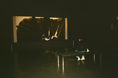 A person sits on a bench in a dimly lit room, observing a large, abstract, monochromatic artwork displayed on the wall. The focus is on the interaction between the viewer and the art piece in a quiet, contemplative setting.