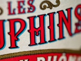 Close-up view of stylized text with bold, vivid red and blue letters outlined in black against a beige textured background. The design includes ornamental scrolls in gold, adding a vintage or classic feel.