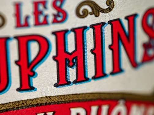 Close-up view of stylized text with bold, vivid red and blue letters outlined in black against a beige textured background. The design includes ornamental scrolls in gold, adding a vintage or classic feel.