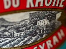 A close-up of a label featuring a detailed illustration of a vineyard hillside with several small buildings. The label includes bold red text with serif fonts.