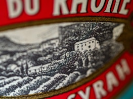 A close-up of a label featuring a detailed illustration of a vineyard hillside with several small buildings. The label includes bold red text with serif fonts.