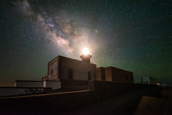 A brightly illuminated lighthouse stands against a backdrop of a vivid starry night sky, featuring the Milky Way galaxy. The lighthouse structure appears solid and angular, with light emanating from its top, casting a glow on the surrounding area.