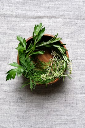 A wooden bowl filled with fresh herbs including rosemary, parsley, and thyme is placed on a textured gray fabric surface.