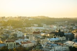 A picturesque cityscape featuring a mix of historical and modern architecture. Prominent buildings with ornate designs stand alongside newer structures. The scene is bathed in warm, golden sunlight, casting a serene glow over the city. Rolling hills with lush greenery frame the background, adding depth to the vista.
